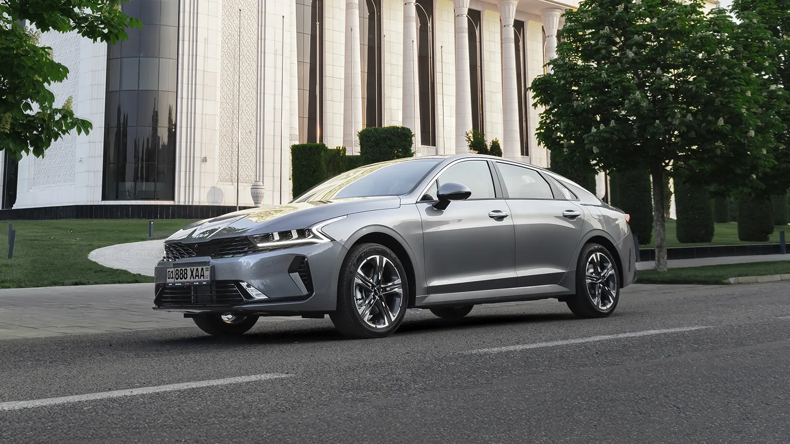 Серый седан Kia K5 спереди сбоку на дороге у здания с белыми колоннами